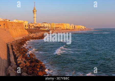 Vista al tramonto della diga e della costa atlantica a Cadice, Spagna, con onde, edifici cittadini e una calda luce dorata lungo il lungomare. Foto Stock