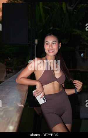 Una giovane donna thailandese felice e atletica marrone sorride mentre tiene il suo smartphone in un balcone all'aperto poco illuminato, che rappresenta un vivace post-lavoro Foto Stock