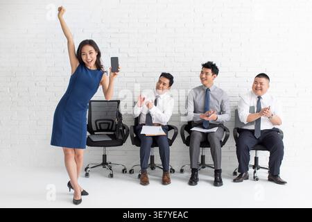 Allegra candidata cinese che solleva il braccio e mostra smartphone mentre viene applaudita da candidati di lavoro di sesso maschile seduti contro il muro di mattoni bianchi ... Foto Stock