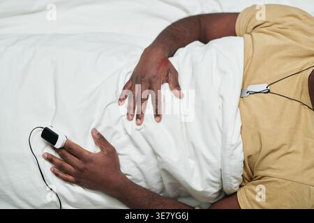 Immagine ritagliata di uomo Nero con pulsossimetro sulla punta del dito giacente sotto un foglio bianco nel letto di ospedale Foto Stock