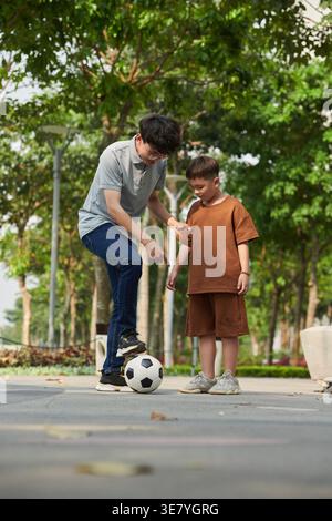 Uomo asiatico che insegna figlio asiatico giocare a calcio mentre si trova in parco durante il giorno Foto Stock