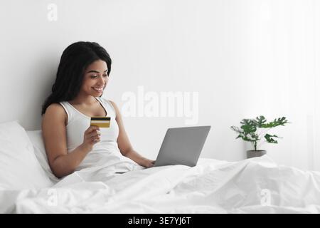 Donna nera sorridente che tiene in mano la carta di credito mentre usa il laptop seduto sul letto coperta sulle gambe in camera da letto a casa Foto Stock