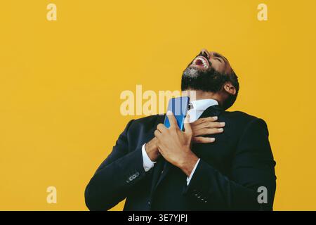 Allegro uomo d'affari barbuto in tuta e cravatta che tiene in mano lo smartphone ridendo con le mani sul petto e la testa gettata indietro mentre si sta in piedi contro... Foto Stock