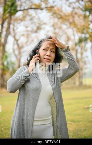 Donna asiatica anziana che guarda lontano toccando la fronte, mentre parla al telefono in piedi sul prato del parco durante il giorno Foto Stock