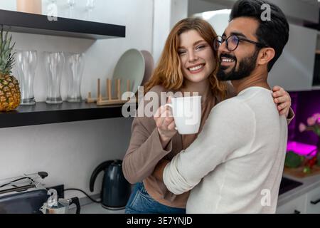 Marito indiano sorridente che indossa occhiali che guardano lontano abbracciando moglie caucasica che tiene una tazza di caffè seduta sul piano della cucina a casa Foto Stock
