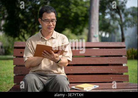Uomo asiatico di mezza età che legge un libro seduto sulla panchina nel parco durante il giorno Foto Stock