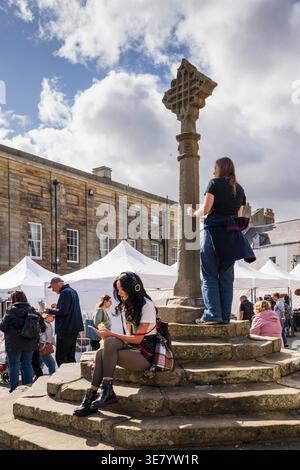 Un vivace mercato all'aperto si snoda tra acquirenti e venditori intorno a uno storico mercato incrociato sotto un cielo parzialmente nuvoloso Foto Stock