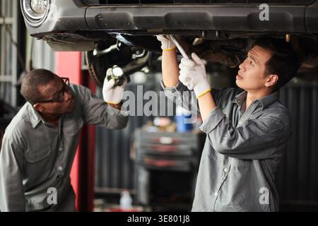 Meccanici maschi neri e asiatici che indossano guanti che riparano sotto il telaio dell'auto mentre si trova in un'officina di riparazione auto Foto Stock