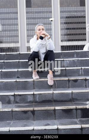 Donna anziana seduta su gradini di pietra nella piazza urbana, indossa felpa con cappuccio, riposa e guarda dopo la pioggia Foto Stock