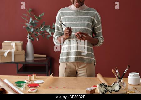 Immagine ritagliata di donna nera in pullover a righe grigie che tiene il pennello e ornamenti natalizi dipingendo con acquerello in piedi a tavola contro... Foto Stock