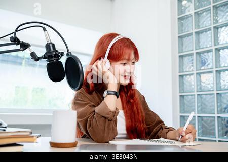 Podcaster donna asiatica che tocca le cuffie tenendo la penna guardando lontano mentre si siede al tavolo con microfono e notebook in casa studio Foto Stock