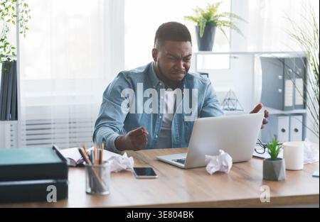 Un uomo d'affari nero stressato che guarda un notebook seduto a un tavolo con pugno chiuso in ufficio Foto Stock