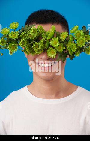 Uomo asiatico sorridente che indossa una camicia bianca con occhi ricoperti di margherite verdi su sfondo blu in studio Foto Stock