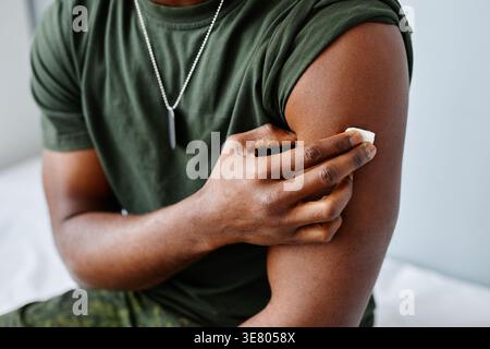 Mano di nero militare uomo che preme cotone su braccio seduto sul letto medico dopo la vaccinazione in clinica Foto Stock