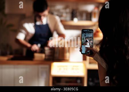 Donna asiatica con lunghi capelli ricci che fotografa un barista che lavora al bancone del bar Foto Stock