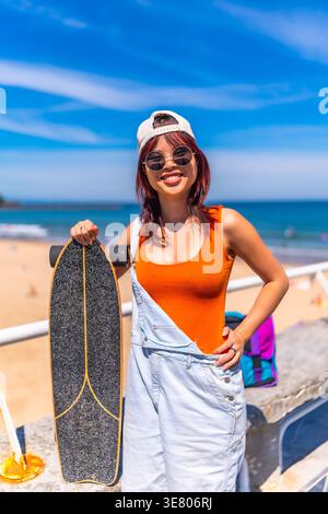 Donna asiatica che indossa berretto e occhiali da sole tenendo lo skateboard in piedi con la mano in vita sulla spiaggia nelle giornate di sole Foto Stock