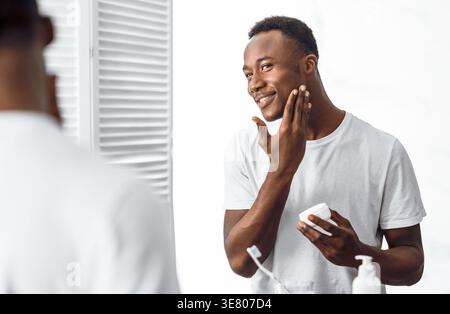 Uomo nero in camicia bianca che regge il vaso idratante e applica la crema sul viso stando in piedi davanti allo specchio nel bagno di casa Foto Stock