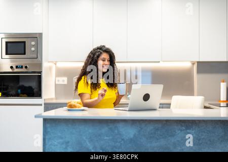 Donna ispanica con capelli ricci che partecipa a una riunione online utilizzando un computer portatile mentre fa colazione al bancone della cucina a casa Foto Stock