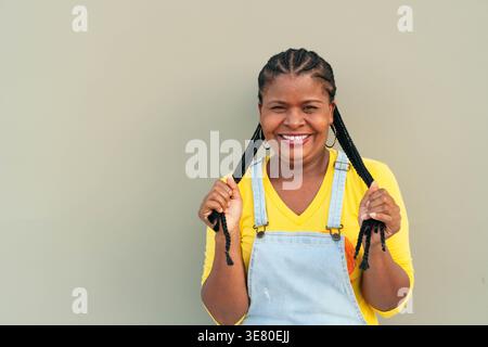 Donna ispanica con trecce che toccano i capelli mentre sta in piedi su sfondo grigio in studio Foto Stock