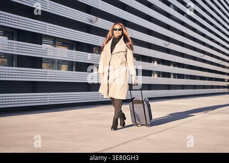 Donna nera che indossa occhiali da sole e cammina con i bagagli fuori dall'edificio dell'ufficio durante il tramonto Foto Stock