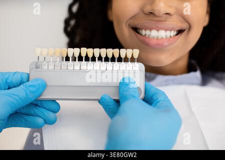 Dentista maschio di colore nero che indossa i guanti che tengono la guida dell'ombreggiatura dentale scelta del colore dei denti per la paziente di sesso femminile in clinica Foto Stock