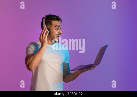 Uomo arabo con barba che indossa una camicia bianca che indossa le cuffie che tengono in mano e guardano lo schermo del laptop mentre si trova in piedi contro il bac rosa e viola... Foto Stock