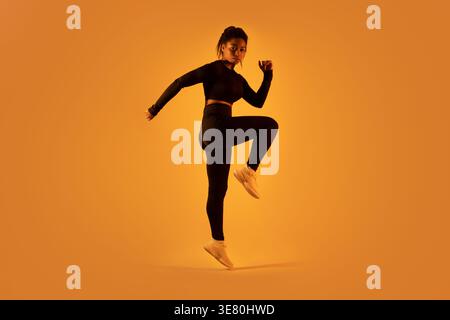 Donna nera che indossa un abbigliamento sportivo che salta sullo sfondo arancione in studio Foto Stock
