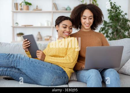 Donna nera che tiene un tablet che guarda un notebook che utilizza un'amica mentre siede sul divano in salotto di casa Foto Stock