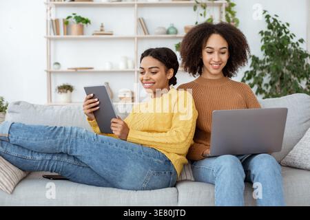 Donna di colore che usa un tablet appoggiato ad un'amica che usa un computer portatile mentre siede sul divano nel salotto di casa Foto Stock