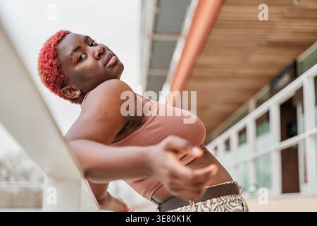 Adolescente afroamericana con i capelli rossi corti in piedi e appoggiata contro ringhiere di metallo sulla strada della città durante il giorno Foto Stock