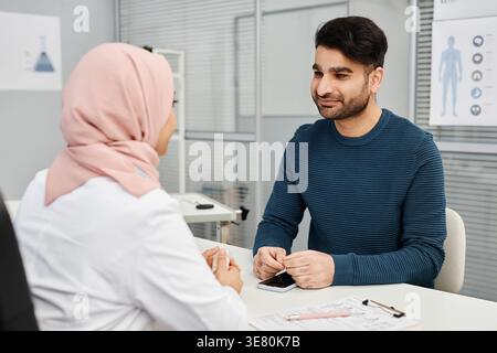 Paziente di sesso maschile di razza mista che parla con un medico donna in hijab mentre è seduto alla scrivania in clinica Foto Stock