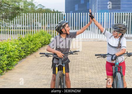 Ciclisti ispanici che indossano caschi che si danno fiocchi alti l'uno all'altro in piedi con le biciclette sul sentiero pedonale del parco durante il giorno Foto Stock