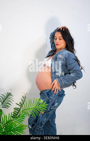 Donna incinta ispanica in piedi con la mano sui capelli e l'anca vicino alle piante in studio Foto Stock