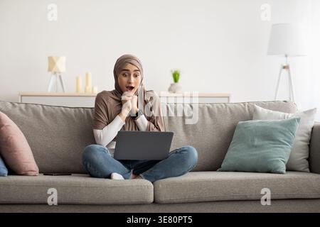 Donna araba scioccata che indossa l'hijab appoggiato al mento su mani strette che guarda lo schermo del notebook mentre è seduta con le gambe incrociate sul divano nel soggiorno di casa Foto Stock