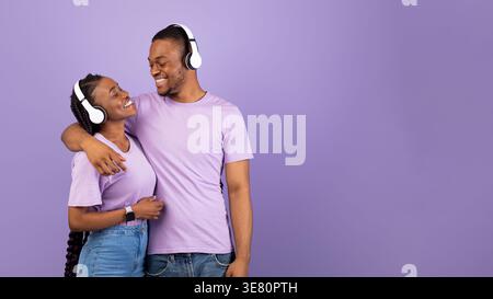 Coppia Smiling Black che abbraccia e ascolta la musica con le cuffie wireless mentre stai in piedi con gli occhi chiusi sullo sfondo viola in studio Foto Stock