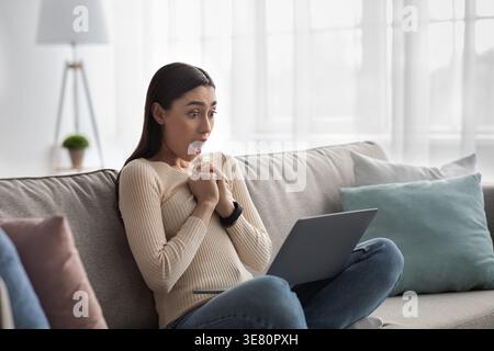 Una donna araba scioccata con le mani strette che guarda lo schermo di un laptop seduto con le gambe incrociate sul divano nel soggiorno di casa Foto Stock