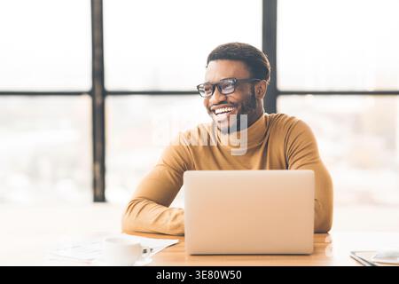 Allegro uomo nero che indossa occhiali da vista che guarda lontano seduto a un tavolo con un computer portatile in ufficio Foto Stock