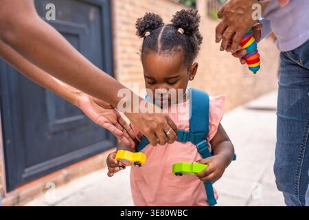 Bambina nera che tiene in mano i giocattoli mentre la madre fissa il suo zaino per strada durante il giorno Foto Stock