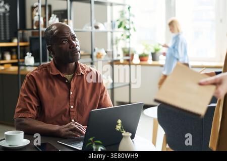 Uomo di mezza età di colore che guarda la cameriera caucasica mentre si siede al tavolo di fronte al computer portatile al bar Foto Stock