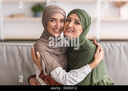 Allegra madre musulmana mediorientale di mezza età in hijab che abbraccia sua figlia mentre siede sul divano nel soggiorno di casa Foto Stock
