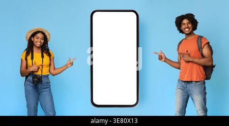 Cheerful Black abbina i turisti con zaini e fotocamera che puntano a uno smartphone enorme mentre si trovano sullo sfondo blu in studio Foto Stock