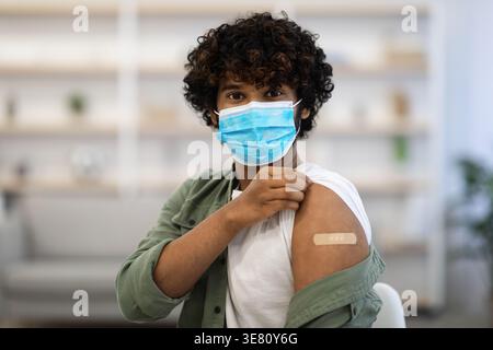 Uomo asiatico con capelli ricci che indossa maschera facciale che mostra bende sul braccio che viene vaccinato mentre seduto sulla sedia in clinica Foto Stock