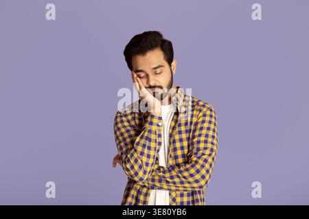 Uomo indiano con la mano sul mento e gli occhi chiusi in piedi su sfondo viola in studio Foto Stock