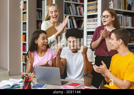 Allegro e variegato gruppo di studenti universitari che festeggiano davanti a un notebook in biblioteca Foto Stock