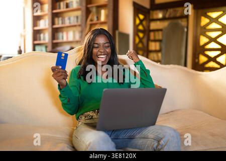 Adolescente di colore con laptop in possesso della carta di credito che celebra il successo seduto sul divano nel soggiorno di casa Foto Stock
