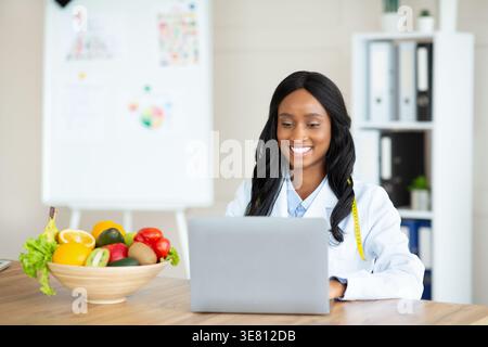 Nutrizionista di colore che lavora su un computer portatile seduto alla scrivania in ufficio Foto Stock