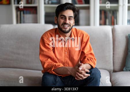 Un uomo sorridente del Medio Oriente con le mani strette seduto sul divano di casa Foto Stock