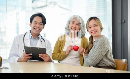 Donna asiatica anziana con capelli bianchi ricci che tengono rosso heLifestyle vicino al petto seduto tra la nipote e il cardiologo maschile seduto a tavola in cl... Foto Stock
