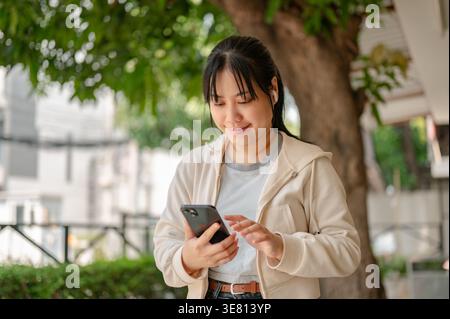 Donna thailandese che indossa auricolari wireless con smartphone in piedi nel parco durante il giorno Foto Stock