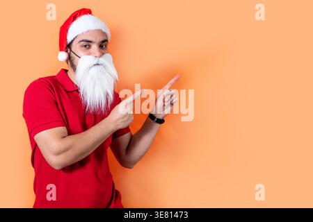 Uomo arabo con cappello di Babbo Natale e barba bianca che punta le dita lateralmente in piedi su sfondo arancione in studio Foto Stock